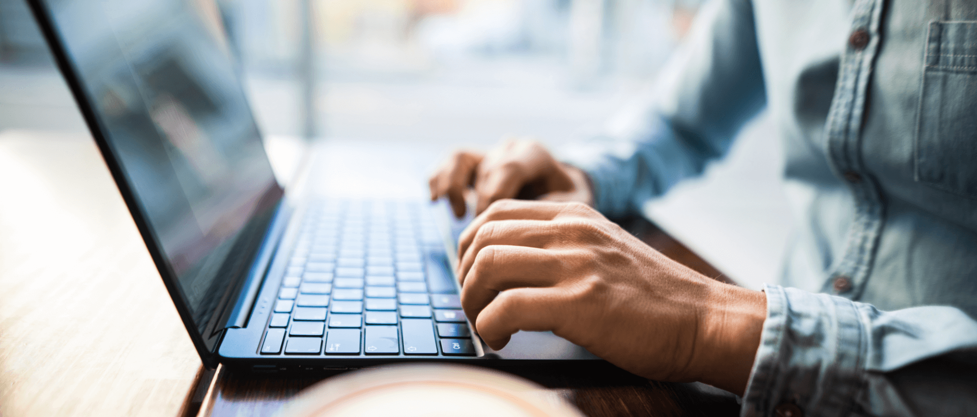 A young man typing on a PEAQ laptop, next to a full coffee cup, sitting in a café, close-up, panorama