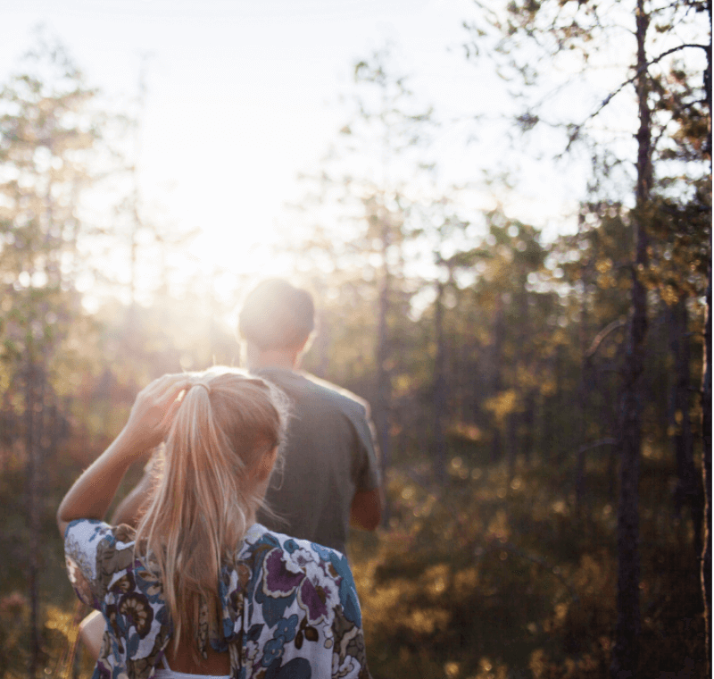 Eine junge Frau folgt einem jungen Mann in den Wald, sommerliche Atmosphäre und Sonneneinfall, Rückansicht