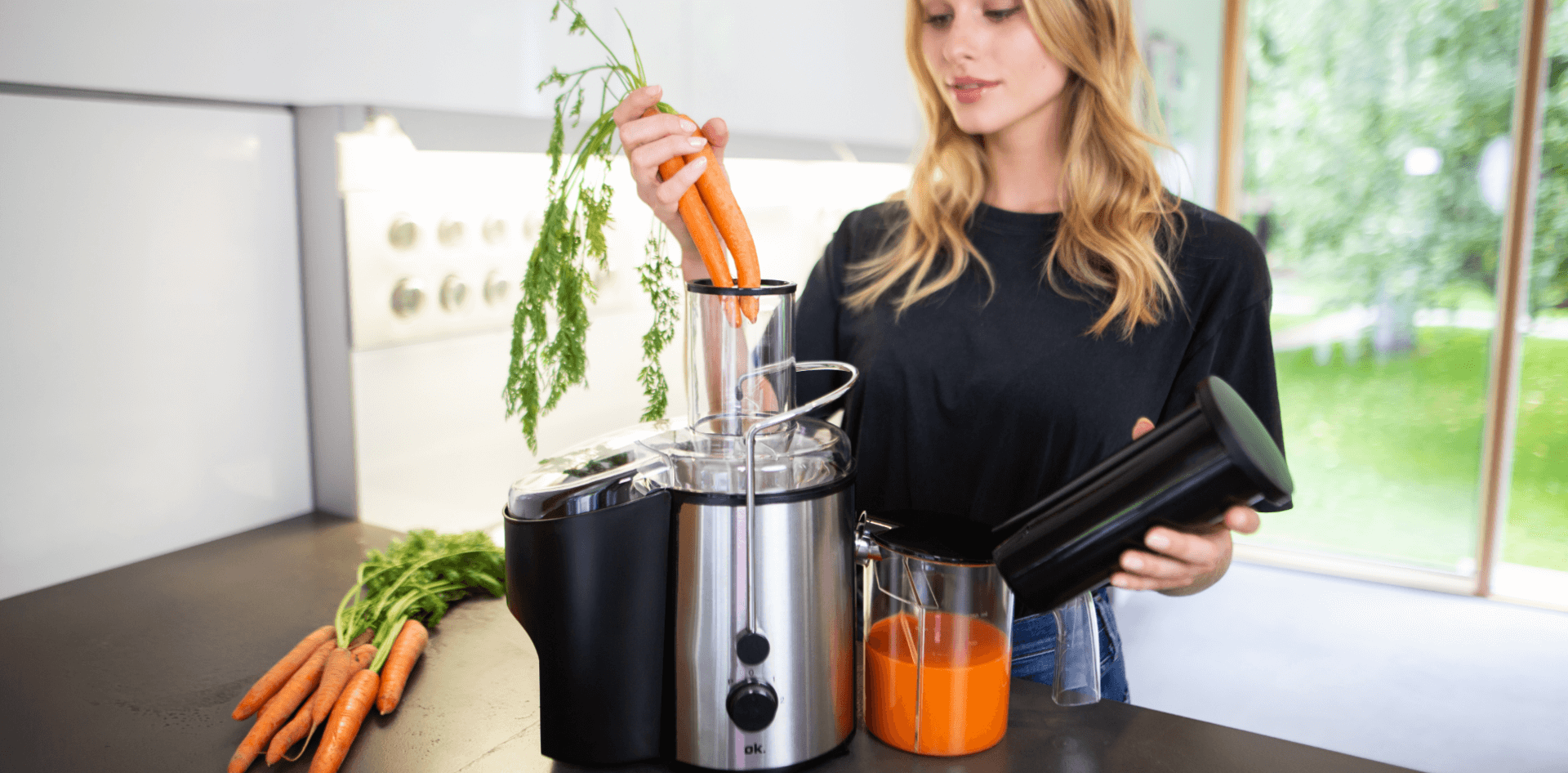 A young woman using an ok. juicer with a bunch of carrots, in a modern kitchen with a view of her garden