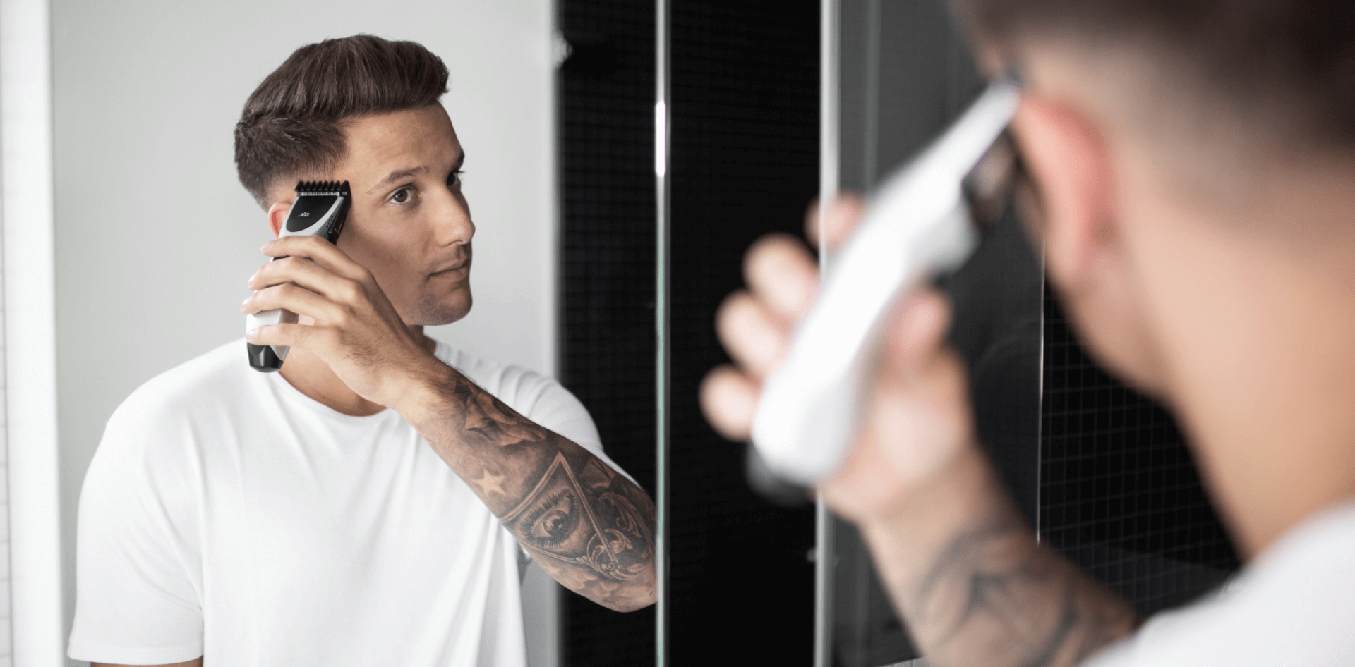 Młody mężczyzna z tatuażem na ręku strzyże przed lustrem włosy maszynką marki ok., nowoczesna łazienka, zbliżenie