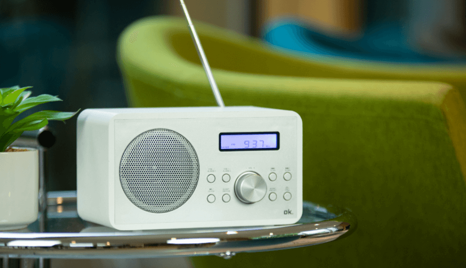 Um rádio+DAB branco da marca ok. de pé sobre uma mesa redonda de vidro numa sala de espera com poltrona verde, vista de perto